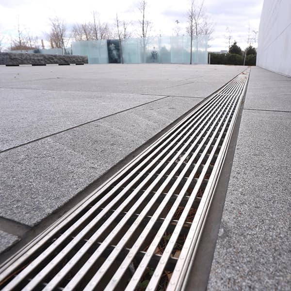 ACO KlassikDrain trench drain with stainless steel grate installed in a commercial plaza, collecting surface water along a paved walkway